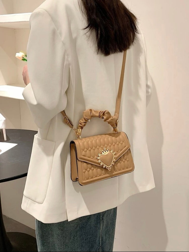 Puede incluir: Un bolso de mano acolchado de color beige con un cierre dorado en forma de coraz&oacute;n y un asa de cuerda trenzada. El bolso tiene una correa larga para llevarlo al hombro.