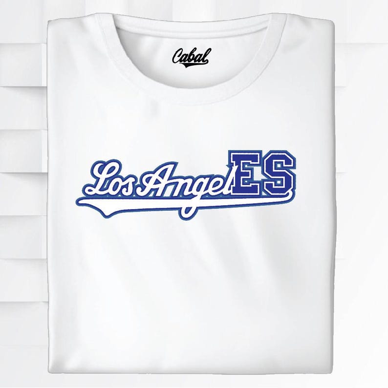 May include: White t-shirt with the text "Los Angeles" in a blue script font, with "ES" in a block letter style. The shirt has a crew neck and the brand name "Cabal" is printed on the collar.