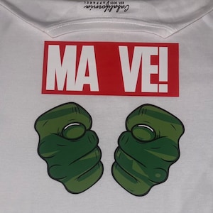 May include: White t-shirt with a red rectangle that says "MA VE!" and two green fists with white knuckles.