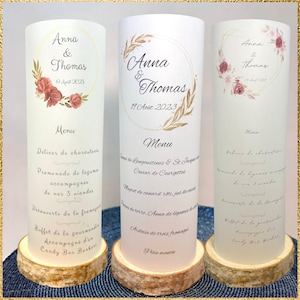 Peut inclure: Trois menus cylindriques blancs portant les prénoms Anna & Thomas et la date du 19 Août 2023. Chaque menu est orné d'illustrations florales et fixé sur un socle circulaire en bois naturel, détaillant les différents plats du repas.