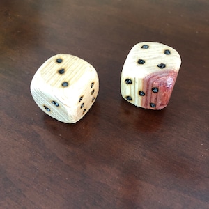 May include: Two wooden dice with black dots. One die is made of light wood and the other is made of a darker wood with a reddish hue.