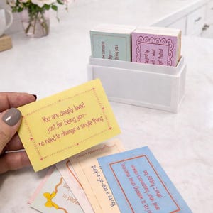 Op de afbeelding: Een witte doos bevat een stapel kleurrijke kaarten met inspirerende boodschappen. Een hand houdt een gele kaart met de tekst "You are deeply loved just for being you - no need to change a single thing." Andere kaarten zijn zichtbaar.