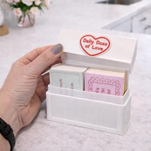 Op de afbeelding: Witte rechthoekige doos met een scharnierend deksel, met het opschrift "Daily Dose of Love" in een rood hart. Binnenin bevinden zich twee stapels kleine kaarten met pastelkleuren en tekst. De doos staat op een wit aanrecht.