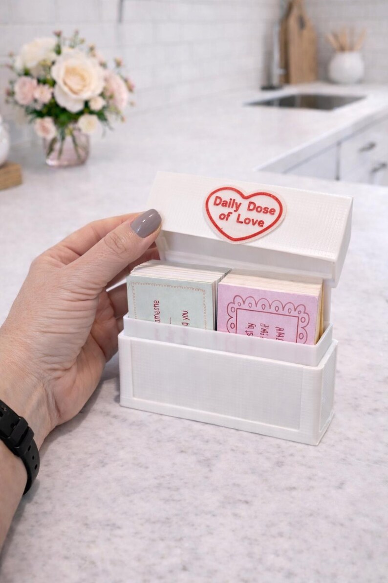 Op de afbeelding: Witte rechthoekige doos met een scharnierend deksel, met het opschrift "Daily Dose of Love" in een rood hart. Binnenin bevinden zich twee stapels kleine kaarten met pastelkleuren en tekst. De doos staat op een wit aanrecht.