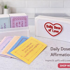 Op de afbeelding: Een witte doos met een rood hartlogo met de tekst "Daily Dose of Love" en een stapel affirmatiekaarten in pastelkleuren. De kaarten bevatten inspirerende boodschappen. De tekst "Daily Dose of Love Affirmation Cards" is ook zichtbaar.