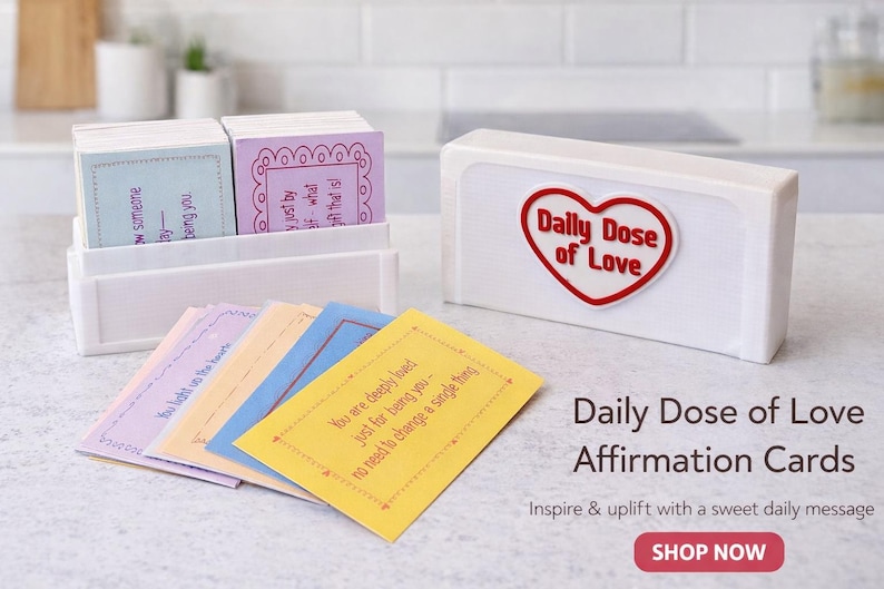 Op de afbeelding: Een witte doos met een rood hartlogo met de tekst "Daily Dose of Love" en een stapel affirmatiekaarten in pastelkleuren. De kaarten bevatten inspirerende boodschappen. De tekst "Daily Dose of Love Affirmation Cards" is ook zichtbaar.