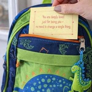 Op de afbeelding: Een blauwgroene rugzak met dinosaurus thema met een gele notitie met de tekst "You are deeply loved just for being you - no need to change a single thing." De rugzak heeft een dinosaurus bedel.