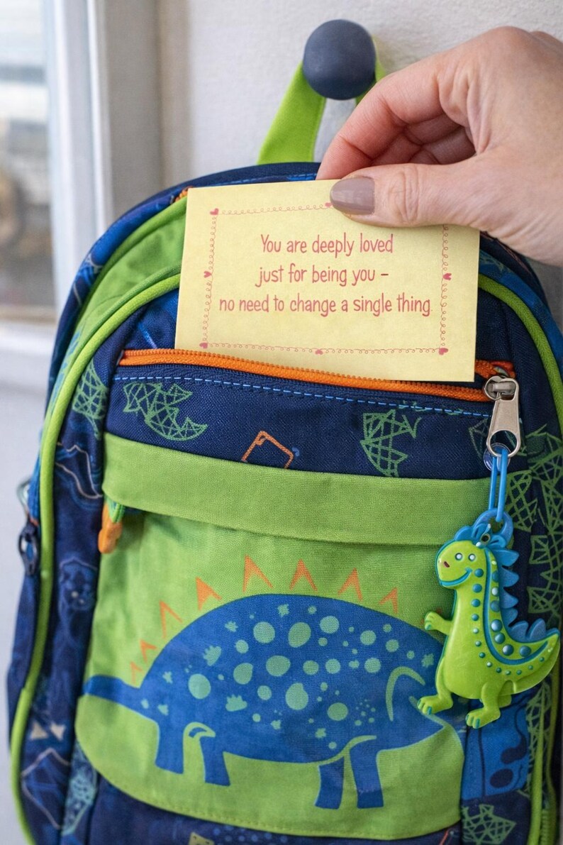 Op de afbeelding: Een blauwgroene rugzak met dinosaurus thema met een gele notitie met de tekst "You are deeply loved just for being you - no need to change a single thing." De rugzak heeft een dinosaurus bedel.