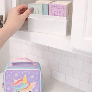 Op de afbeelding: Een witte rechthoekige container met kleurrijke kaarten, met een hand die erin reikt om er een te selecteren. Daaronder staat een paarse en blauwe eenhoorn lunchtas met een sterrenpatroon en een regenboogmanen op een wit aanrecht.