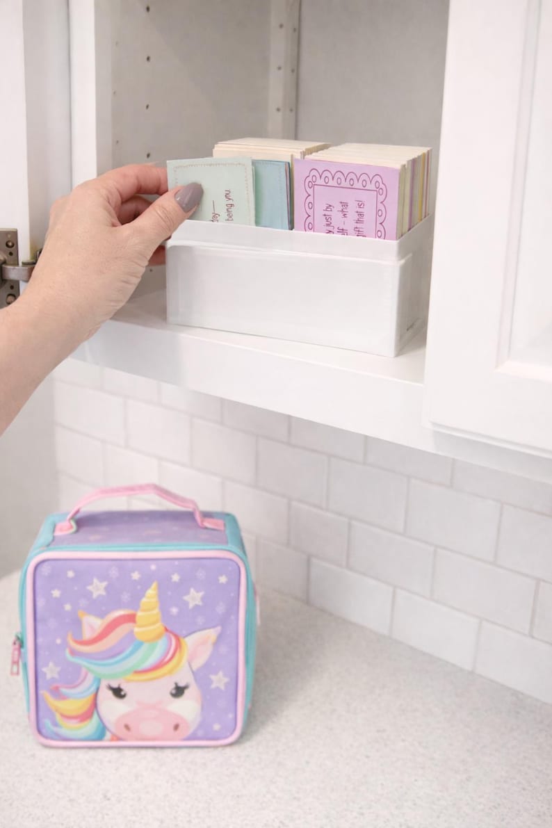 Op de afbeelding: Een witte rechthoekige container met kleurrijke kaarten, met een hand die erin reikt om er een te selecteren. Daaronder staat een paarse en blauwe eenhoorn lunchtas met een sterrenpatroon en een regenboogmanen op een wit aanrecht.