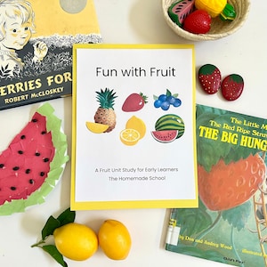 May include: A yellow card with the text "Fun with Fruit" and a fruit unit study for early learners. The card is on a white surface with a watermelon craft, a book about blueberries, and a book about a hungry bear.