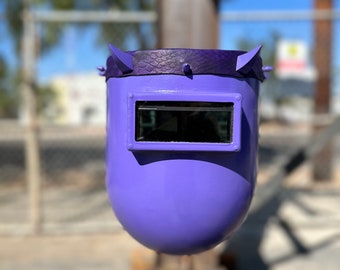 May include: A purple welding helmet with a black visor and two small horns on top. The helmet is hanging from a metal beam.