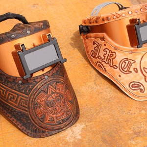 May include: Two copper-coloured welding helmets with leather straps and decorative engravings. One helmet has a skull and crossbones design with the text "J.R.C." The other helmet has a circular design with a stylised animal face and a Greek key pattern.