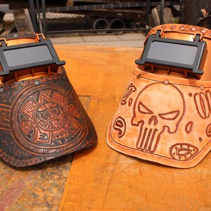May include: Two leather welding helmets with different designs. One helmet features a skull and crossbones design, while the other has an Aztec-inspired design. Both helmets are brown and have a copper-coloured metal frame.