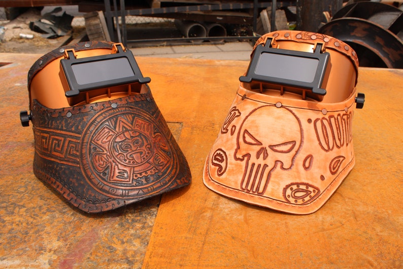 May include: Two leather welding helmets with different designs. One helmet features a skull and crossbones design, while the other has an Aztec-inspired design. Both helmets are brown and have a copper-coloured metal frame.
