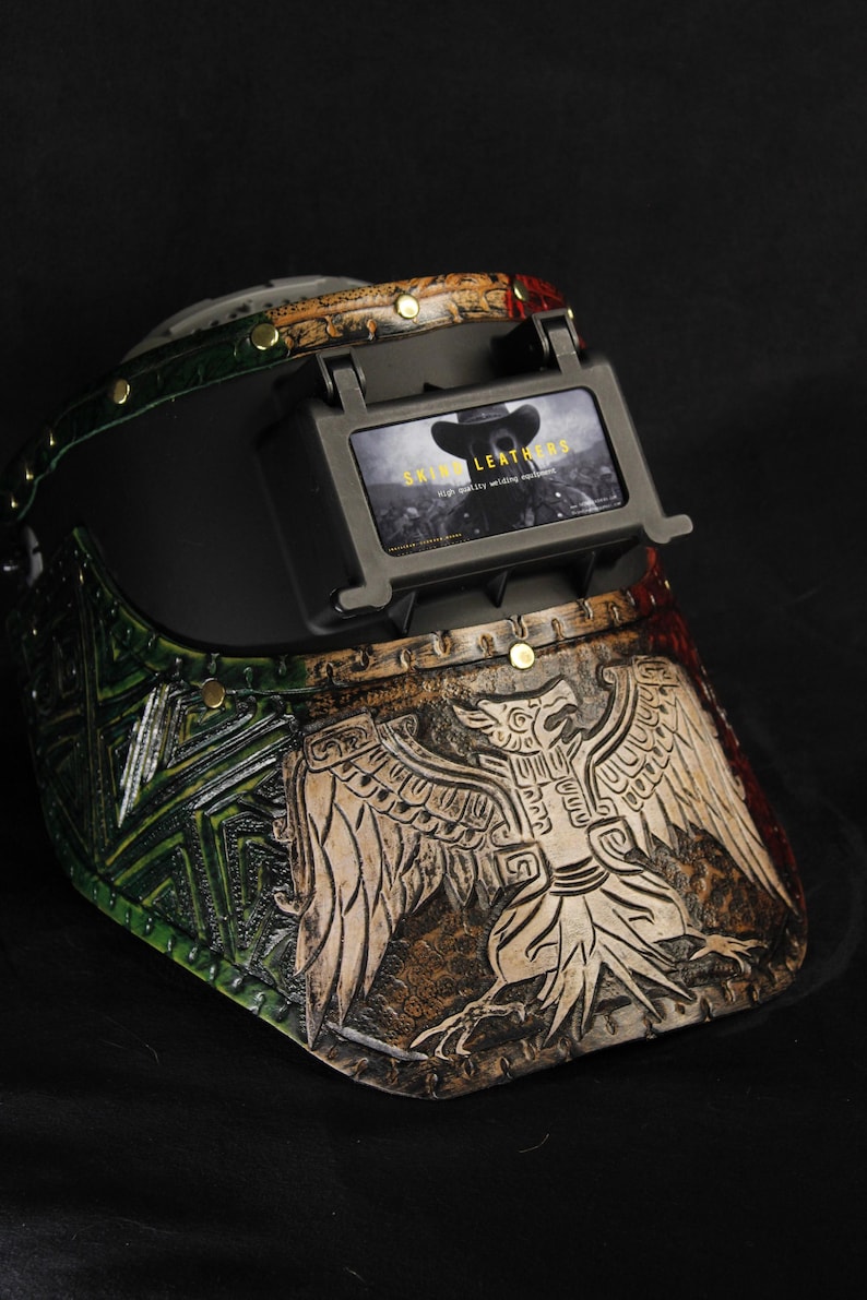 Op de afbeelding: Op maat gemaakte leren lashelm met een adelaarsontwerp. De helm heeft een gedetailleerd, handgemaakt adelaarsmotief in bruin en beige, met groene en rode accenten. De tekst "SKIN LEATHERS" is zichtbaar op het scherm.