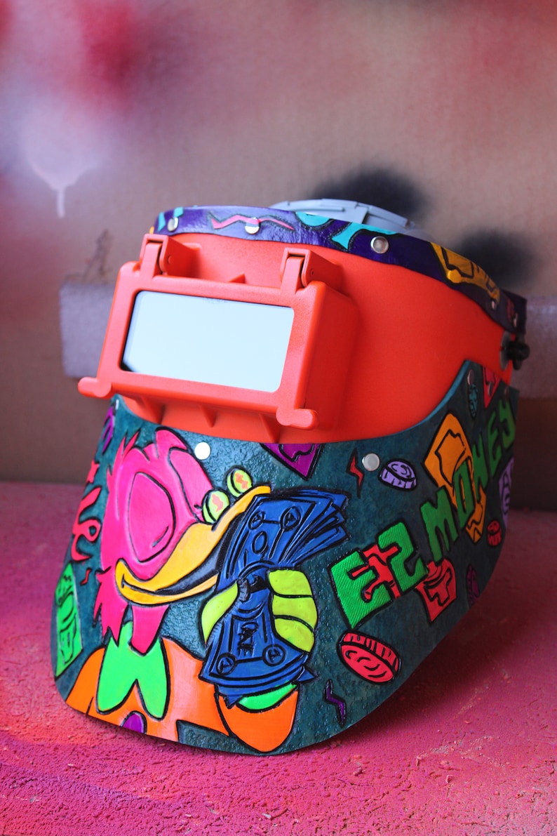 May include: A bright orange welding mask with a clear visor. The mask is decorated with a colorful cartoon character holding money and the words "EZ MONEY".