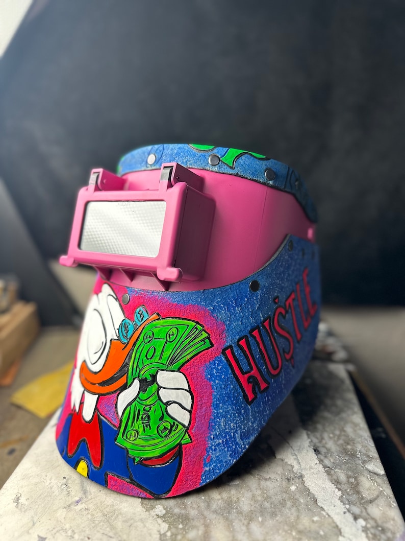 May include: A pink welding helmet with a blue and green design. The helmet has a cartoon image of a duck holding a stack of money. The text "Hustle" is written on the side of the helmet.