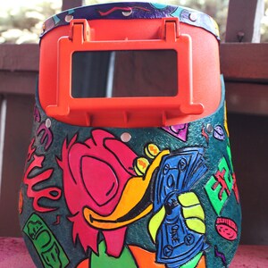 May include: A bright orange welding helmet with a green, blue, and yellow cartoon illustration of a duck holding money. The duck is wearing a green bow tie and has a pink beak. The helmet has a clear viewing window.