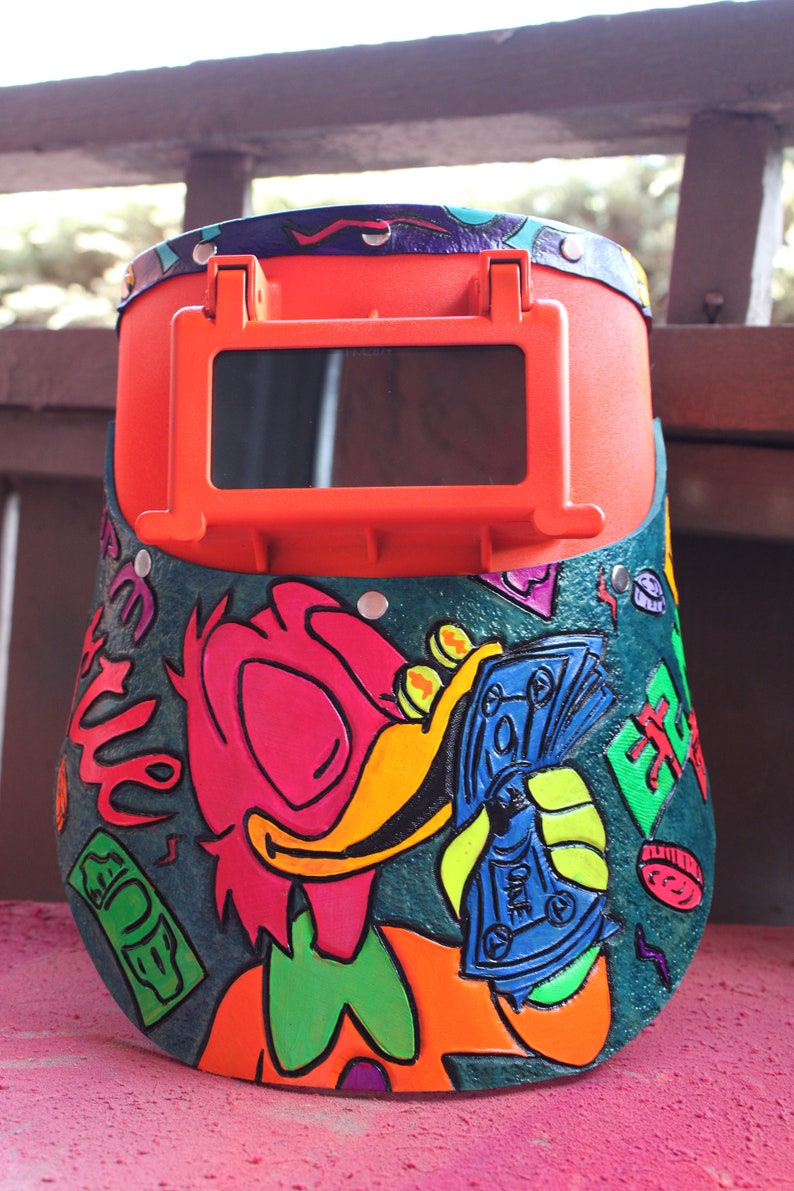 May include: A bright orange welding helmet with a green, blue, and yellow cartoon illustration of a duck holding money. The duck is wearing a green bow tie and has a pink beak. The helmet has a clear viewing window.