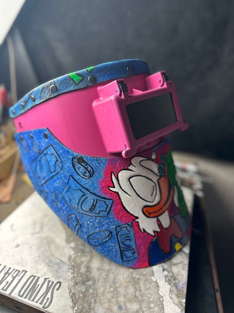 May include: A pink welding helmet with a blue and green cartoon design featuring a duck character and money bills. The helmet has a dark tinted visor.
