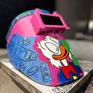 May include: A pink welding helmet with a clear visor and a cartoon illustration of a duck wearing a blue suit and holding money. The helmet is decorated with a blue and white money pattern.