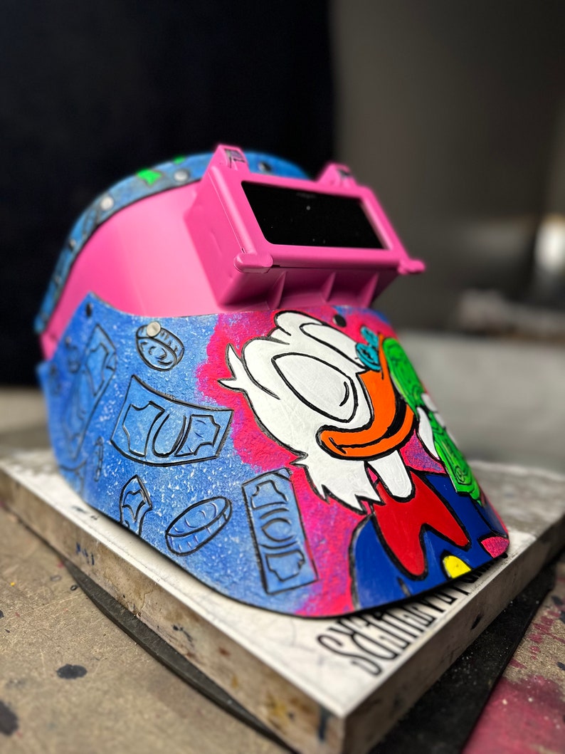 May include: A pink welding helmet with a clear visor and a cartoon illustration of a duck wearing a blue suit and holding money. The helmet is decorated with a blue and white money pattern.