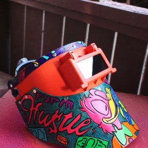 May include: A bright orange welding helmet with a clear visor. The helmet is decorated with a colorful design that includes the word "Hustle" and dollar bills.