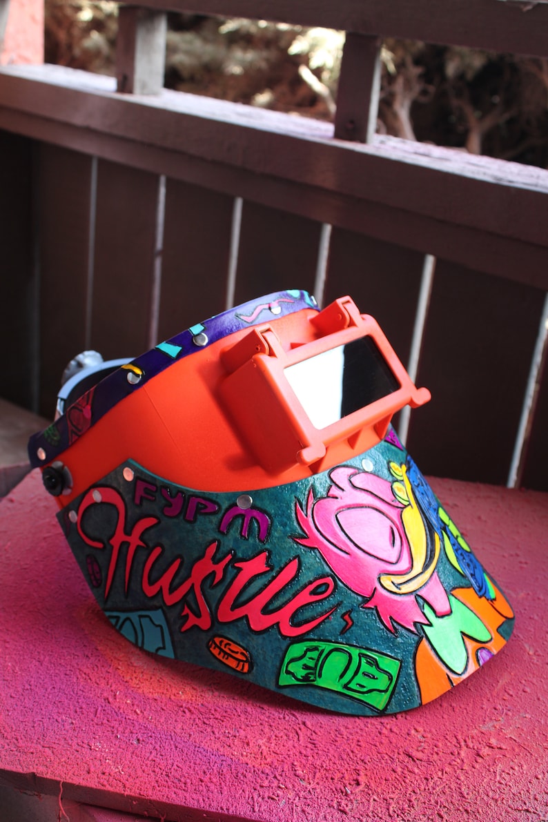 May include: A bright orange welding helmet with a clear visor. The helmet is decorated with a colorful design that includes the word "Hustle" and dollar bills.
