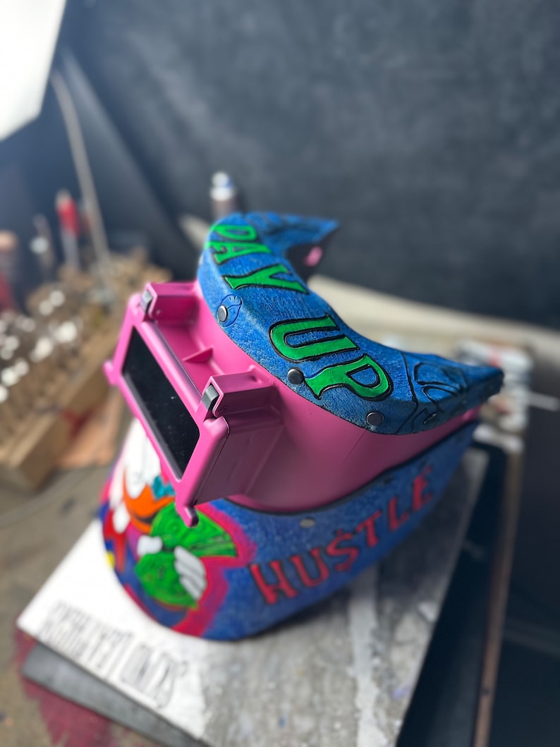 May include: A pink welding helmet with a blue and green design. The helmet has a clear visor and the words "Pay Up" and "Hustle" are written on it.