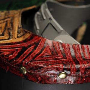 Op de afbeelding: Close-up van een decoratief leren object met een geometrisch patroon. Het object heeft een combinatie van rode, zwarte en gouden kleuren. Goudkleurige studs zijn zichtbaar langs de rand van het object. Het object lijkt deel uit te maken van een groter stuk.