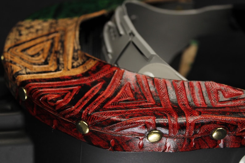 Op de afbeelding: Close-up van een decoratief leren object met een geometrisch patroon. Het object heeft een combinatie van rode, zwarte en gouden kleuren. Goudkleurige studs zijn zichtbaar langs de rand van het object. Het object lijkt deel uit te maken van een groter stuk.