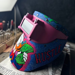 May include: A pink welding helmet with a blue, green, and white design. The helmet has a cartoon character holding money and the word "HUSTLE" written on it.