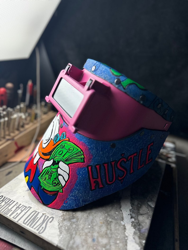May include: A pink welding helmet with a blue, green, and white design. The helmet has a cartoon character holding money and the word "HUSTLE" written on it.