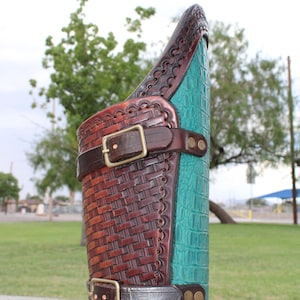 May include: A brown leather arm guard with a woven pattern and a teal alligator-embossed leather panel. The guard has a brass buckle and two straps.