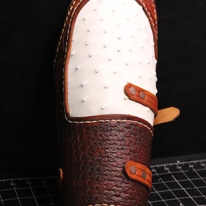 May include: A brown leather arm guard with white ostrich skin and brown leather straps with brass buckles.