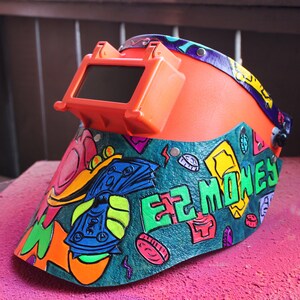 May include: A bright orange welding helmet with a dark tinted visor. The helmet is decorated with a colorful design featuring money bills, coins, and the words "EZ MONEY".