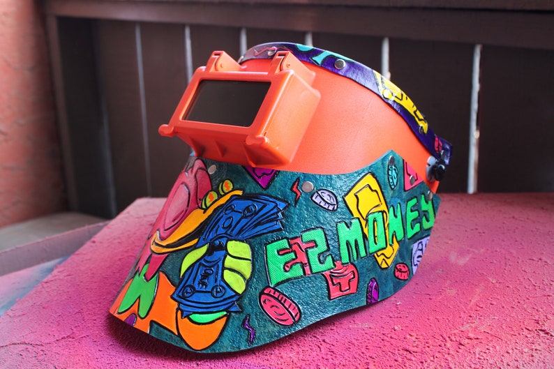 May include: A bright orange welding helmet with a dark tinted visor. The helmet is decorated with a colorful design featuring money bills, coins, and the words "EZ MONEY".