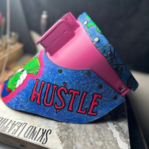 May include: A pink welding helmet with a blue and green design. The helmet has the word "HUSTLE" written in red letters on the front.