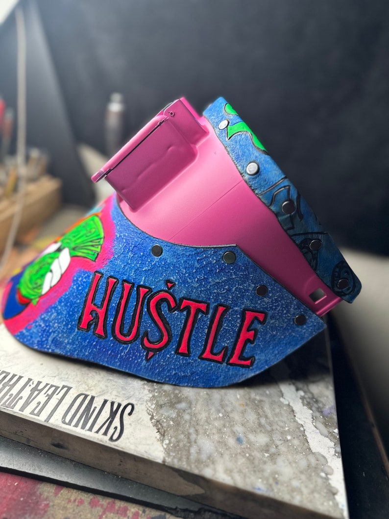 May include: A pink welding helmet with a blue and green design. The helmet has the word "HUSTLE" written in red letters on the front.