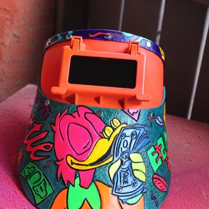May include: A bright orange welding helmet with a dark visor. The helmet is decorated with a cartoon duck holding money, and the words "Get Rich" and "Club" are written on the side.