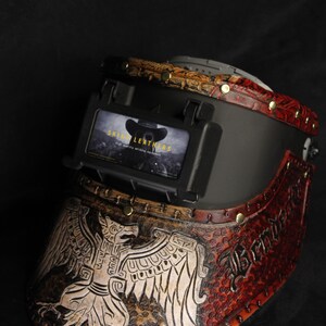Op de afbeelding: Op maat gemaakte lashelm met een gedetailleerd adelaarsontwerp en de tekst "Skinny Leathers". De helm heeft bruine, rode en zwarte lederen accenten met messing klinknagels. Een uniek stuk voor lassers.