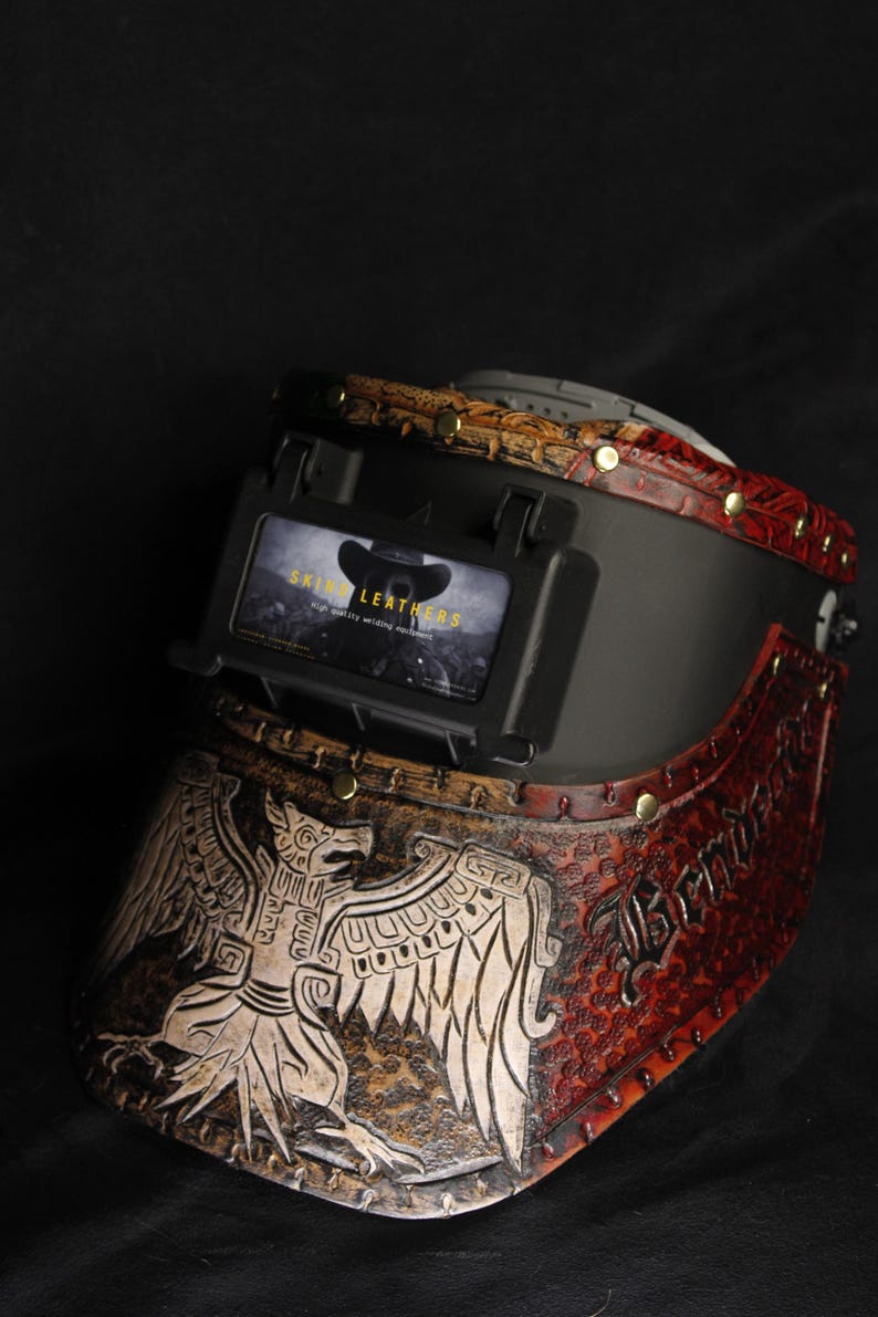Op de afbeelding: Op maat gemaakte lashelm met een gedetailleerd adelaarsontwerp en de tekst "Skinny Leathers". De helm heeft bruine, rode en zwarte lederen accenten met messing klinknagels. Een uniek stuk voor lassers.