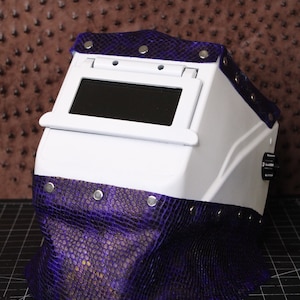 May include: White welding helmet with a purple leather flap and a dark tinted visor. The helmet has a knob on the side and is decorated with silver rivets.