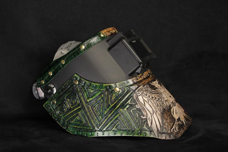 Op de afbeelding: Een op maat gemaakte lashelm met een groen-bruin kleurenschema. De helm heeft ingewikkelde geometrische en vogelontwerpen, messing klinknagels en een donkere lens. De algehele esthetiek is een mix van vakmanschap en functionaliteit.