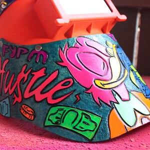May include: A bright orange welding helmet with a green, blue, and pink cartoon character design. The helmet has a clear visor and the words "FYPM Hustle" are painted on the side.