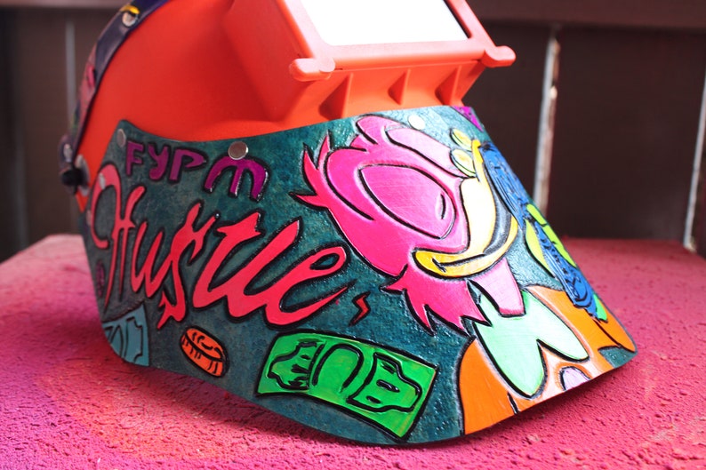 May include: A bright orange welding helmet with a green, blue, and pink cartoon character design. The helmet has a clear visor and the words "FYPM Hustle" are painted on the side.