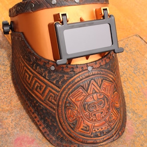 May include: A brown leather welding helmet with a gold-coloured metal frame and a black viewing window. The helmet has a decorative design with a circular pattern and a stylised face.