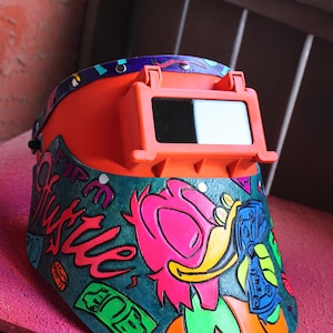 May include: A bright orange welding helmet with a clear visor. The helmet is decorated with a colorful cartoon character and the words "Fyp Hustle" in a graffiti style.