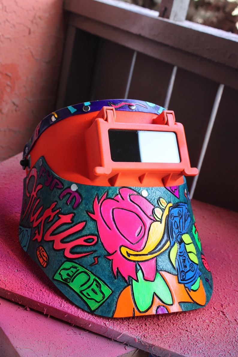 May include: A bright orange welding helmet with a clear visor. The helmet is decorated with a colorful cartoon character and the words "Fyp Hustle" in a graffiti style.